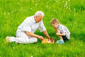 La imagen muestra a un anciano con cabello canoso y anteojos jugando ajedrez con su nieto en un parque. El niño, con una gorra azul, se concentra en su siguiente movimiento. Ambos están sentados sobre una manta en el pasto, rodeados de árboles. La imagen transmite una sensación de alegría, intergeneracionalidad y conexión.